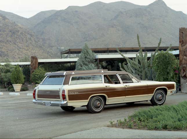 A 1970 Ford LTD Country Squire station wagon in cream and brown parked outside of a modernist house with mountains in the background 