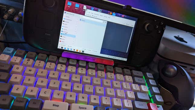 A Steam Deck sits on a dock in front of a keyboard and trackball mouse.