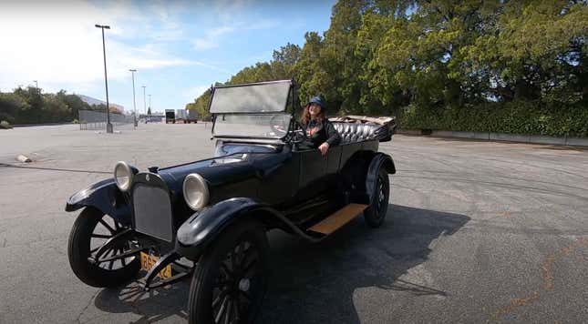 A curly-haired woman in a bucket hat sits smugly in a black 1915 Dodge Bros Touring Car in the parking lot.
