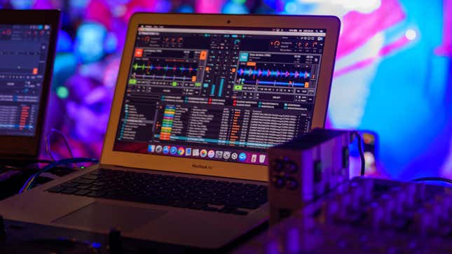 A macBook displaying an audio mixing program sits on a table next to other audio equipment in a night club atmosphere