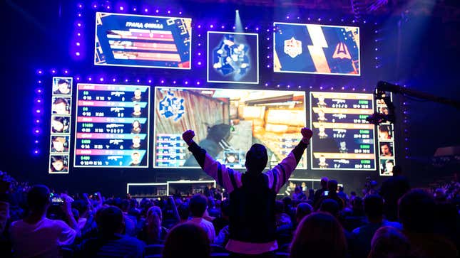 A fan stands up in a crowd at an esports event, his arms wide in celebration before giant screens.