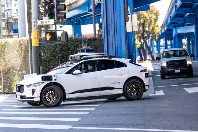 A Waymo self-driving car making a right hand turn in San Francisco. There's a person wearing a ball cap in the driver's seat and bright blue pylons in the background.  