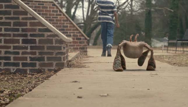 A small alien walks behind a young man on a sidewalk