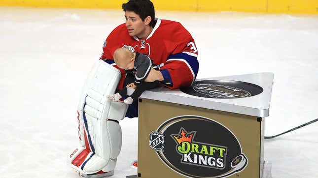 A hockey goalie holding a baby next to a box with the Draft Kings logo on its side 