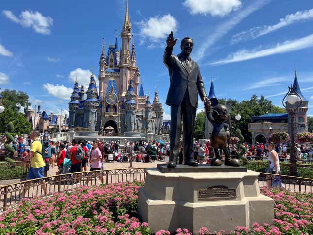 A statue of Walt Disney waving sits on a plinth surrounded by pink flowers. The Disney World castle rises in the distance, while visitors walk around on a sunny day.