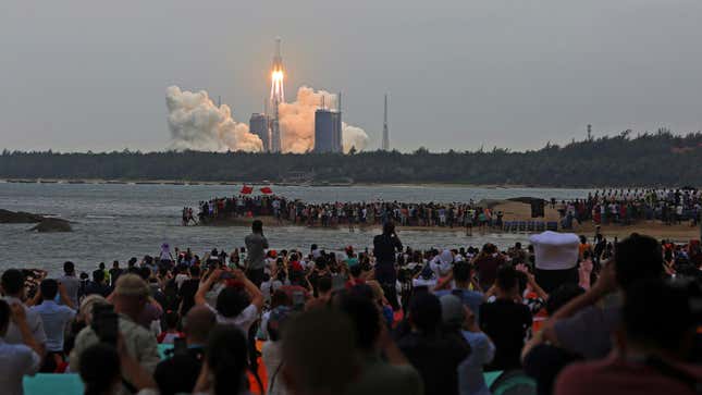 A crowd of people stand on the shore to watch a rocketship blast office from its station