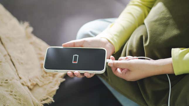 A woman sits on the floor plugging a charging cord into a smartphone displaying a low battery icon
