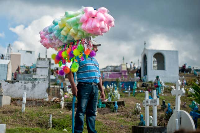 A man holds a stick with bags of cotton candy and fuzzy toys balanced on it. He's standing in a cemetery.