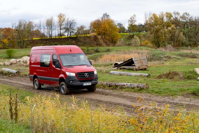 a red 2023 mercedes benz sprinter awd van driving on a dirt trail through a field, with autumn trees and rolling hills in the background