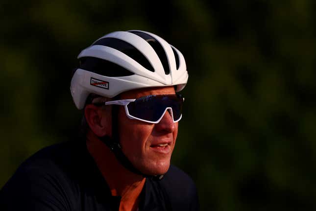 A man in a cycling helmet and goggles squints into the sun while a sheen of sweat appears on his face.