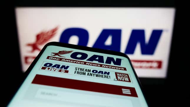A phone with OAN One America News App held close to the camera across from a blurred TV screen with OAN on it