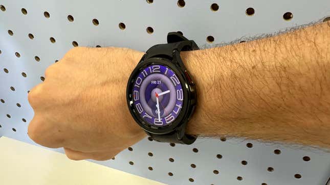 The Galaxy Watch6 Classic on a hairy arm with a purple watch face on a corkboard background.