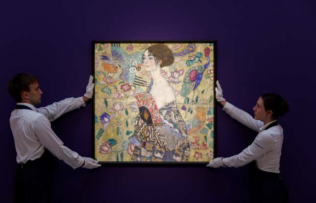 Two people dressed in white shirts and wearing white gloves hold up Gustav Klimt's "Lady with a Fan" against a dark purple wall.