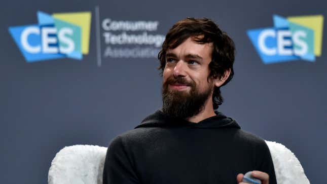 Jack Dorsey sitting in front of a wall that says CES staring off and smiling with a microphone in his hand.