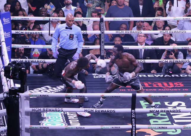 Inside a boxing ring surrounded by a crowd, a Black man in black shorts and gray gloves leans towards his opponent, who wears black shorts and white gloves, while a ref looks on.