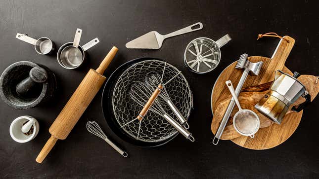 Top-down photo of assorted kitchen tools (mortars and pestles, a wooden rolling pin, metal measuring cups, pie servers, whisks, colanders, wooden cutting boards, stovetop espresso pot, small mesh strainer, flour sifter, and a meat tenderizer) on a matte black surface.