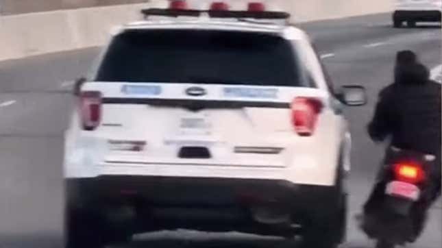 A NYPD cruiser swerves into the same lane as a moped rider on a highway. 