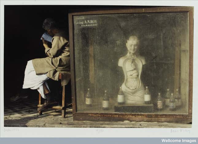 Colour photograph showing an anatomical model advertising a pharmacy in Delhi, India. The model is behind glass and surrounded with bottles. The pharmacist’s name, Kaviraj A N Roy, BSc, MASFRMP, is written on a label at the top left of the glass case. A man, perhaps the pharmacist, sits reading a book. Photograph, 1993. By Ben Dray.