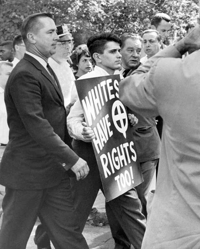 Demonstrator John Patler, carrying sign, is surrounded after his arrest by Englewood, N.J., police during anti-segregation demonstration in Englewood’s McKay Park on August 18, 1962. Patler was one of the several demonstrators from right-wing group calling itself the American National Party. Patler’s was the only arrest during the rally. 