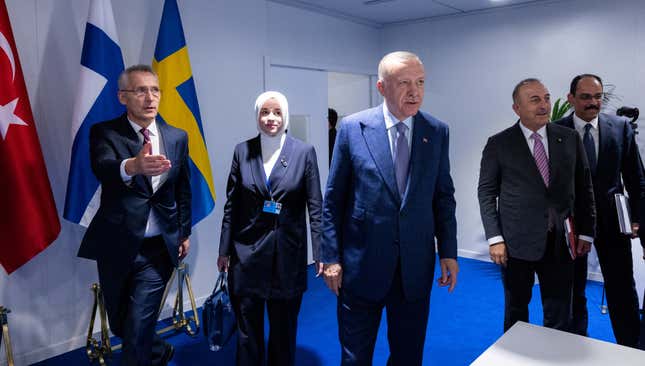 NATO Secretary General Jens Stoltenberg with his hand out, Turkish President Tayyip Erdogan, and three other people, all looking off camera and wearing suits. Various flags are against the wall of what appears to be a conference room with a very blue floor.