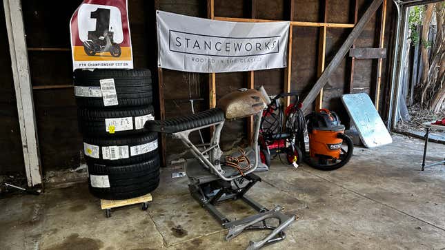 A motorcycle frame is sitting on a lift in front of a big banner that says Stanceworks.