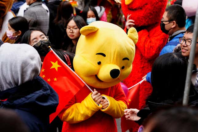 A person wears a Winnie the Pooh costume while holding a Chinese flag. They stand in a crowd of people.