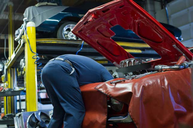 A mechanic in blue coveralls is bent over the hood of a car.