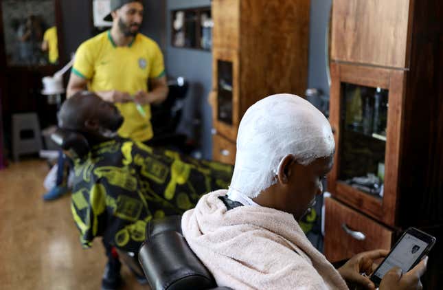 A man with shaving cream on his head looks at his phone while he waits for the barber.