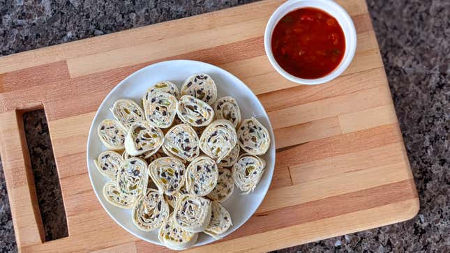 A white plate of Cheesy Tortilla Roll-Ups, or pinwheels, beside a cup of salsa on a wooden cutting board