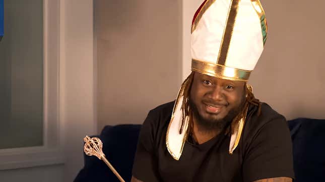 T-Pain wearing a pope hat and cepter while looking down the barrel of a camera lens 