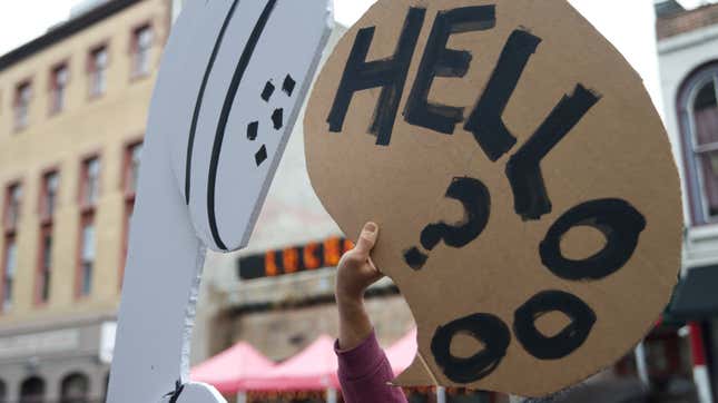 A hand holds up a cardboard sign displaying a telephone receiver speech bubble with the word "Hello?" 