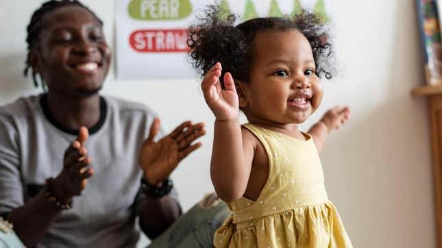 Toddler walking, father sitting behind her and clapping 