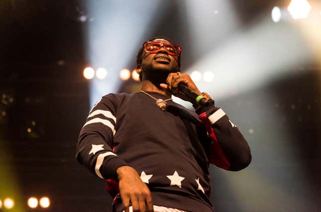 Gucci Mane stands on stage with a microphone wearing a black shirt with white stars and large gold pendant.