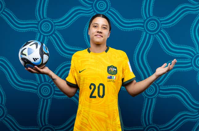 A woman with dark hair in a ponytail, wearing a yellow jersey, shrugs to the camera while holding a soccer ball in one hand before a blue background.