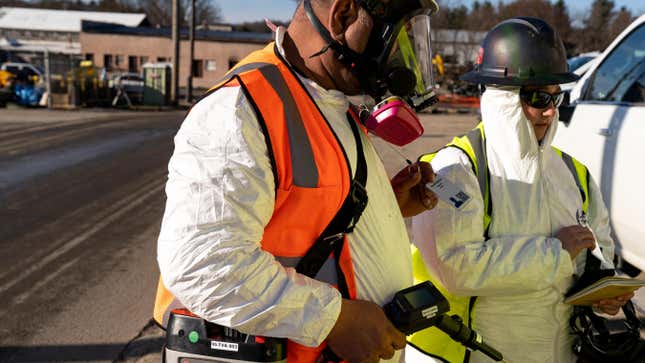 Two people in white chemical suits wearing reflective vests. One, a black man in the foreground, wears a gas mask and holds a testing device in his hand. His white, female counterpart wears only sunglasses and holds a notebook. 