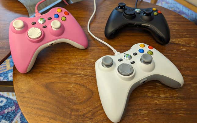 Three Xbox 360 replica controllers, one pink, one white, and one black, sit on a wooden table.