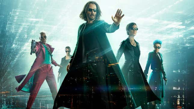 Keanu Reeves, wearing sunglasses and a long black coat, poses with his hand out in front of the the Matrix Resurrections cast.