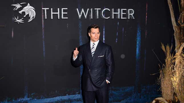 Henry Cavill stands in front of a sign for The Witcher at a red carpet event. 