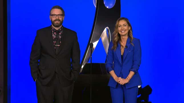 A screenshot from the 2020 DICE Awards depicting hosts Jessica Chobot and Greg Miller on stage against a very blue background.