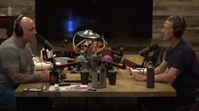 Joe Rogan in headset sits in front of a microphone sits across from Mark Zuckerberg with identical setup.