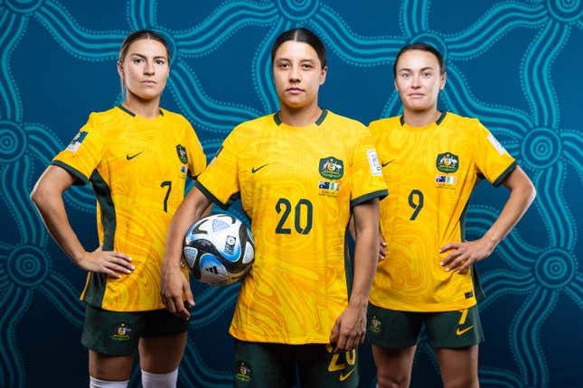 Three white women with dark brown hair pose for a photo while wearing matching yellow tops and green shots. The woman in the middle holds a soccer ball under her arm.