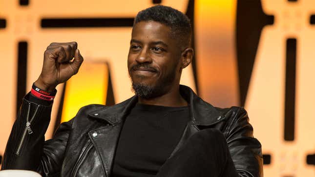 Ahmed Best sits on a stage while holding his fist in the air and smiling. 