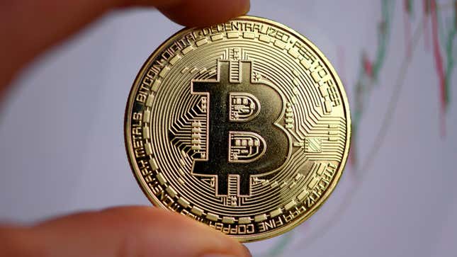 A hand holds a physical representation of a Bitcoin with a stock chart visible in the background
