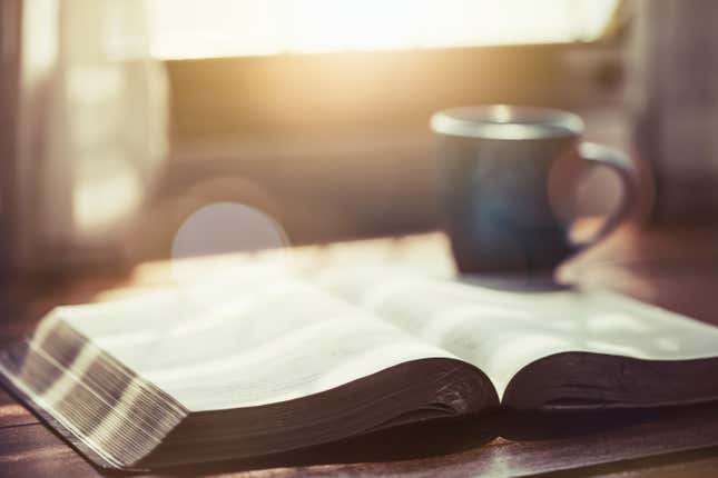 A close up of an open Bible with a cup of coffee for morning devotion on a wooden table with window light