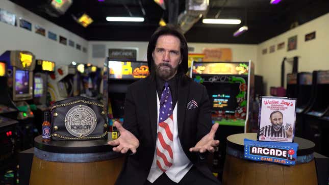Billy Mitchell, wearing a black suit with an American flag tie, sites in front of two Donkey Kong barrels and a bunch of arcade cabinets.
