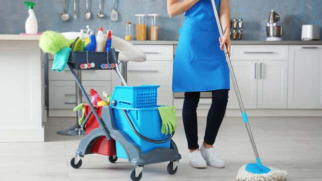 A person wearing jeans, white sneakers, and a blue apron mops a kitchen floor. There's a cart next to them with cleaning supplies and a bucket. 