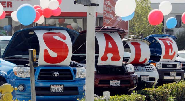 a photo of cars lined up outside a car dealership on a sunny day. four cars have posters hung on their hoods spelling out "sale", and red white and blue balloons are tied to the cars