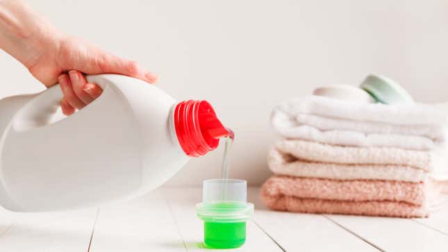 Close-up shot of a person pouring green laundry detergent into a clear cap. There's a pile of folded pink and white towels in the background. 