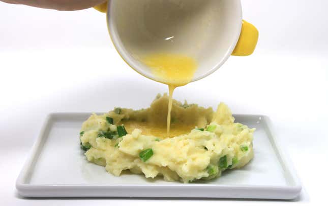 Bowl of butter being drizzled onto pile of mashed potatoes mixed with scallions