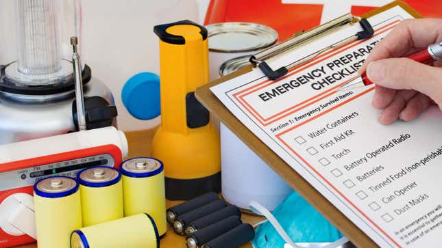 A person checks items off an emergency preparedness checklist. In the background there are batteries, a flashlight, face masks, a radio, and canned food.